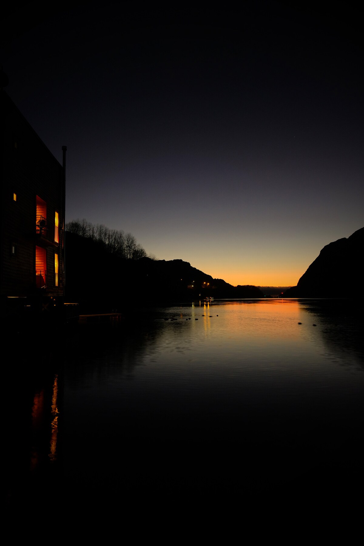 A tranquil evening scene captures a sunset over still waters, with the reflection of warm light from the house contrasting against the darkening sky. Silhouettes of mountains frame the view, enhancing the peaceful atmosphere of the surroundings.