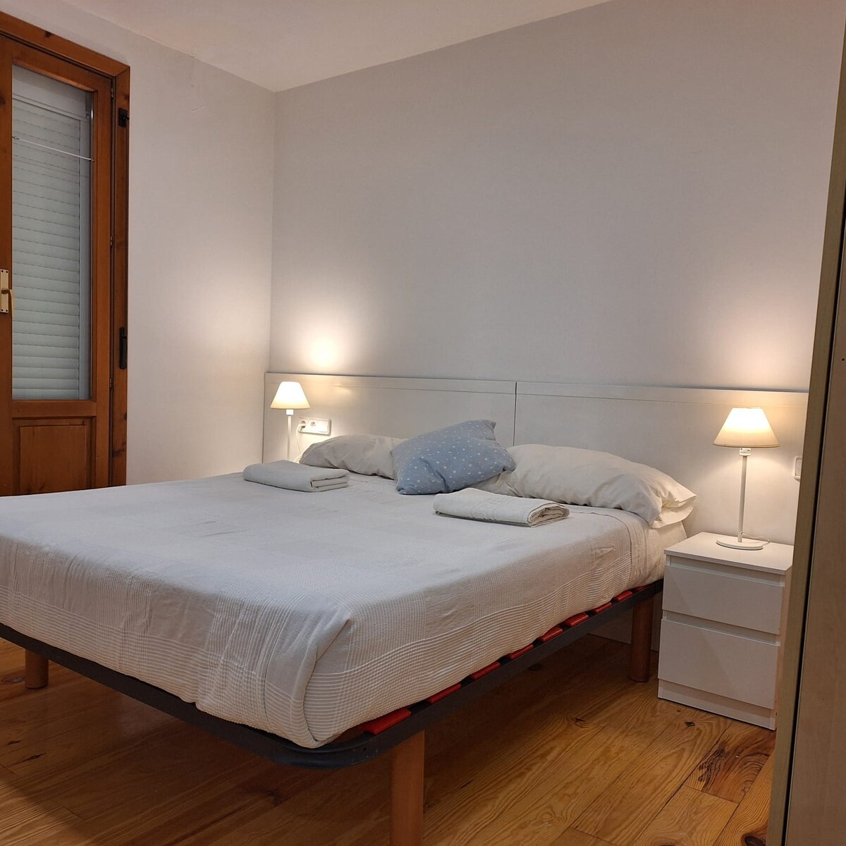 A bright sleeping area features a spacious bed with neatly arranged bedding and two bedside lamps casting soft light. A light-coloured wall complements the natural wood tones of the floor, creating a calming ambiance.