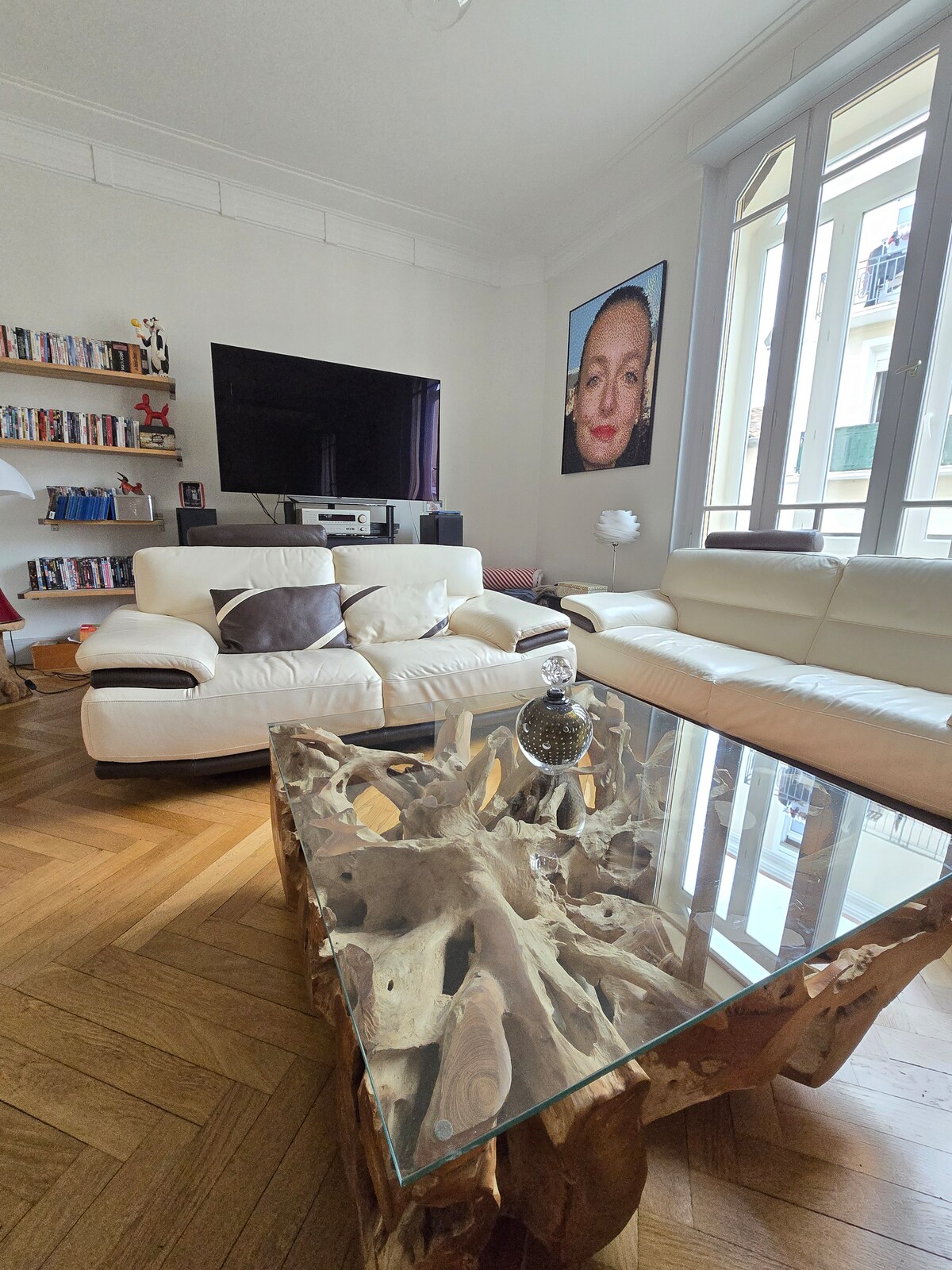 A spacious living area features a large white sectional sofa with decorative cushions, complemented by a distinctive wooden coffee table with a glass top. Natural light filters through large windows, illuminating the room. A shelf lined with books and a television enhance the cozy ambiance.