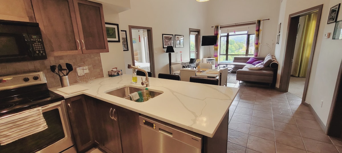The image captures a spacious kitchen area featuring modern appliances, including a stainless steel stove and dishwasher. A marble countertop extends to create a pass-through to the living area, which includes a cozy seating arrangement and large windows offering views of the exterior landscape.