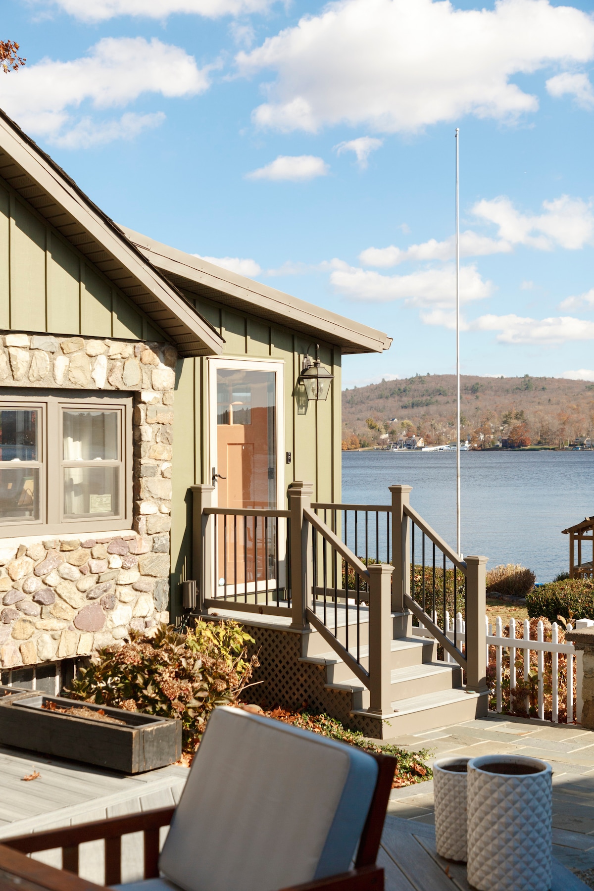 The entrance to the cabin features a wooden deck with a handrail leading to a door. Stone accents are seen on the exterior walls, surrounded by greenery. The view displays the lake in the background, with gentle waves and a clear blue sky above.
