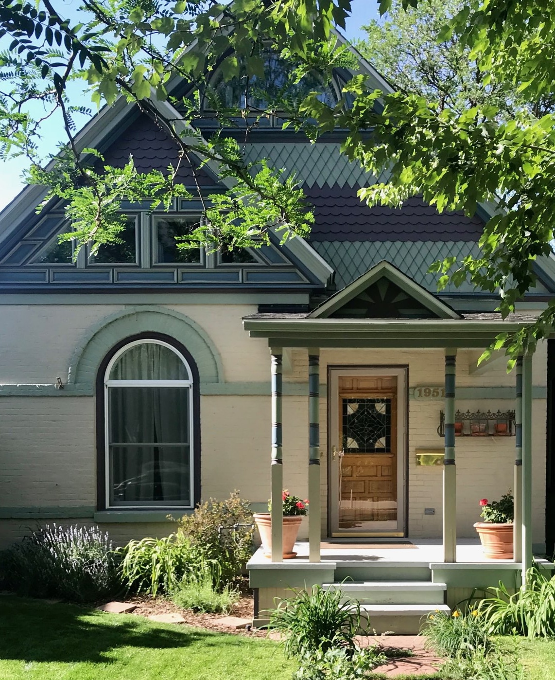 The exterior of a classic Victorian home is showcased, featuring a charming porch with potted flowers. Elaborate architectural details are highlighted by the sun, while a large window allows natural light to illuminate the entryway. Lush greenery surrounds the property, enhancing the inviting atmosphere.