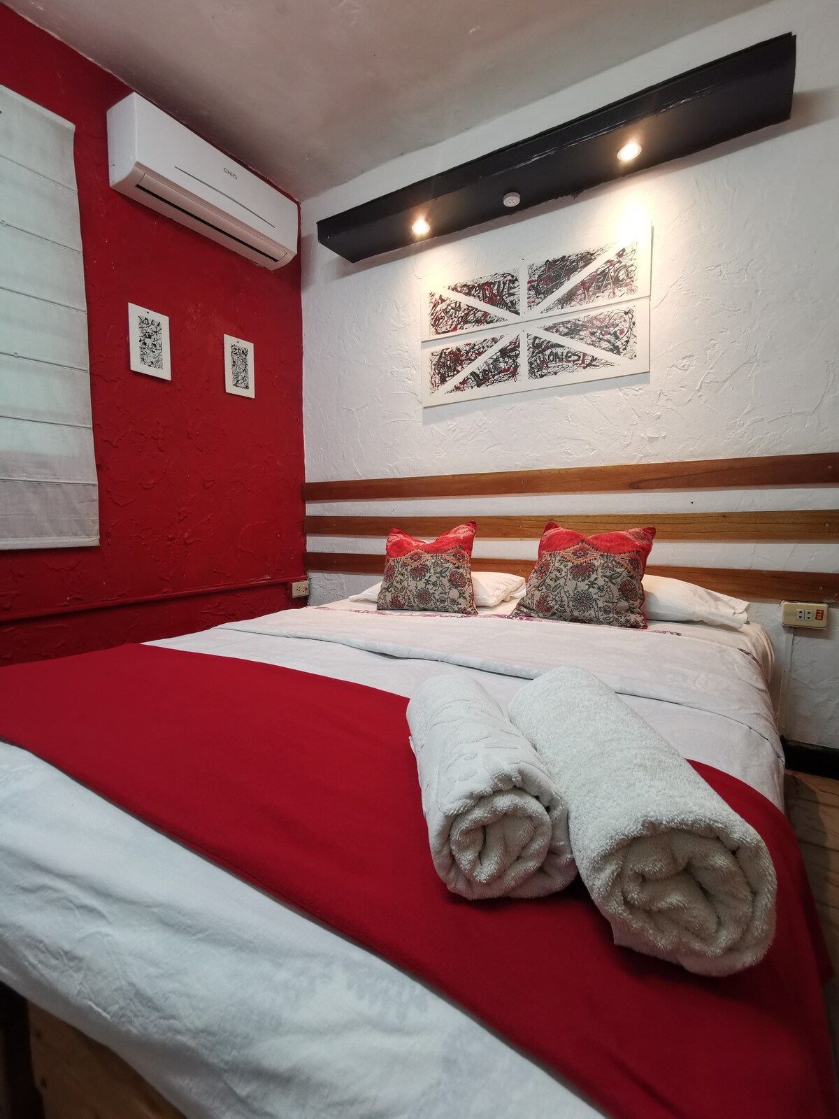 A comfortable sleeping area is highlighted by a spacious bed dressed in crisp white linens and a red accent blanket. Two decorative pillows add a touch of color. The walls are adorned with artwork, while soft lighting illuminates the cozy space.