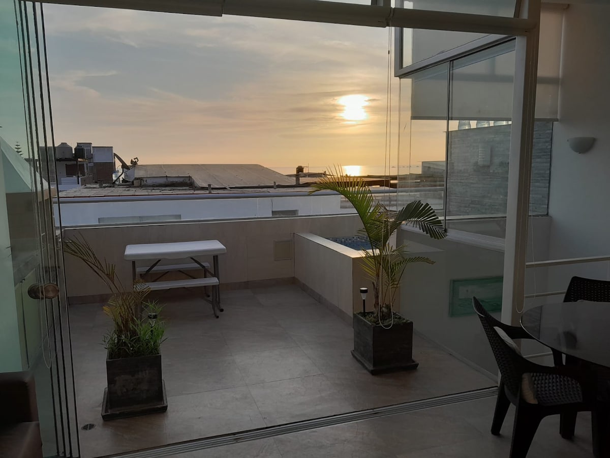 A spacious terrace features a view of the sunset over the sea, framed by large glass panels. Potted plants add a touch of greenery, while a table and chairs provide a setting for relaxation. The tiled floor enhances the open and inviting space.