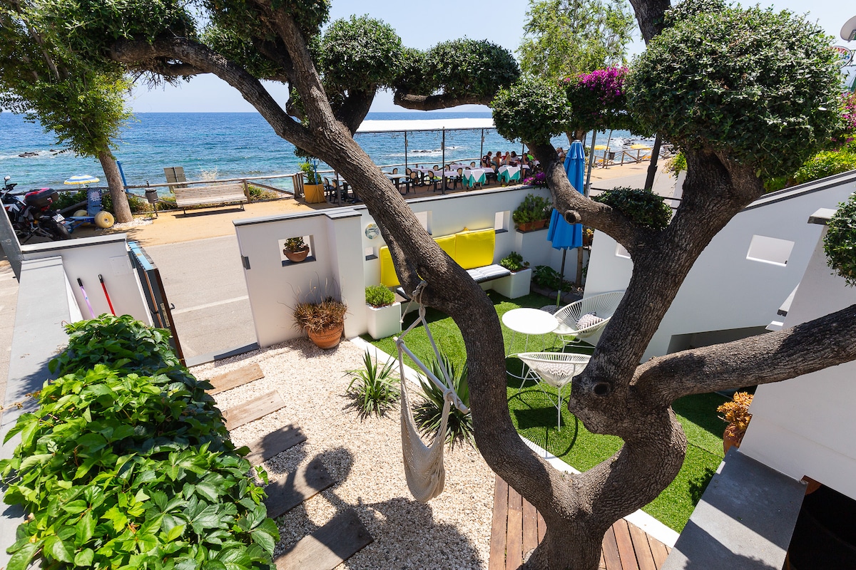 A private outdoor space features a well-maintained garden with a tree providing shade. White furniture is arranged on a small deck, and a hammock is visible. The sea can be seen in the background, with a restaurant situated along the shoreline.