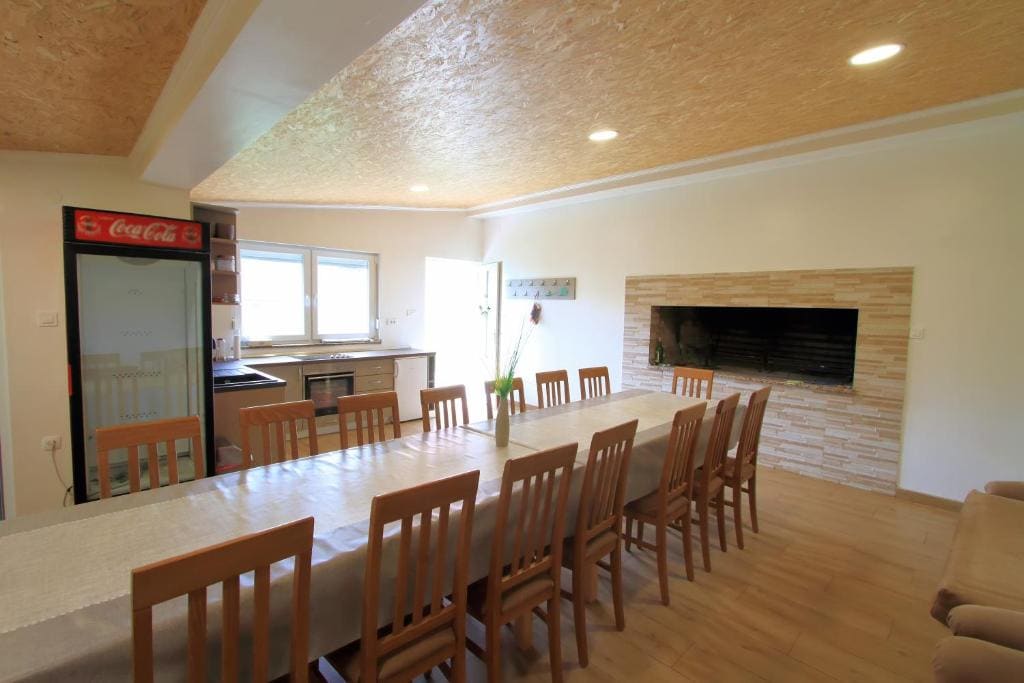 A spacious dining area is presented with a long table surrounded by wooden chairs, ideal for gatherings. A stone fireplace is visible along the wall, while the adjacent kitchenette features modern appliances. Large windows allow natural light to brighten the room.