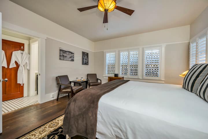 Cama tamaño king con sábanas blancas, manta marrón y dos almohadas a rayas. Dos mesas auxiliares y lámparas. Arte mural en blanco y negro. Persianas. Dos sillones de cuero marrón, mesa de centro, ventilador de techo y vista al baño.