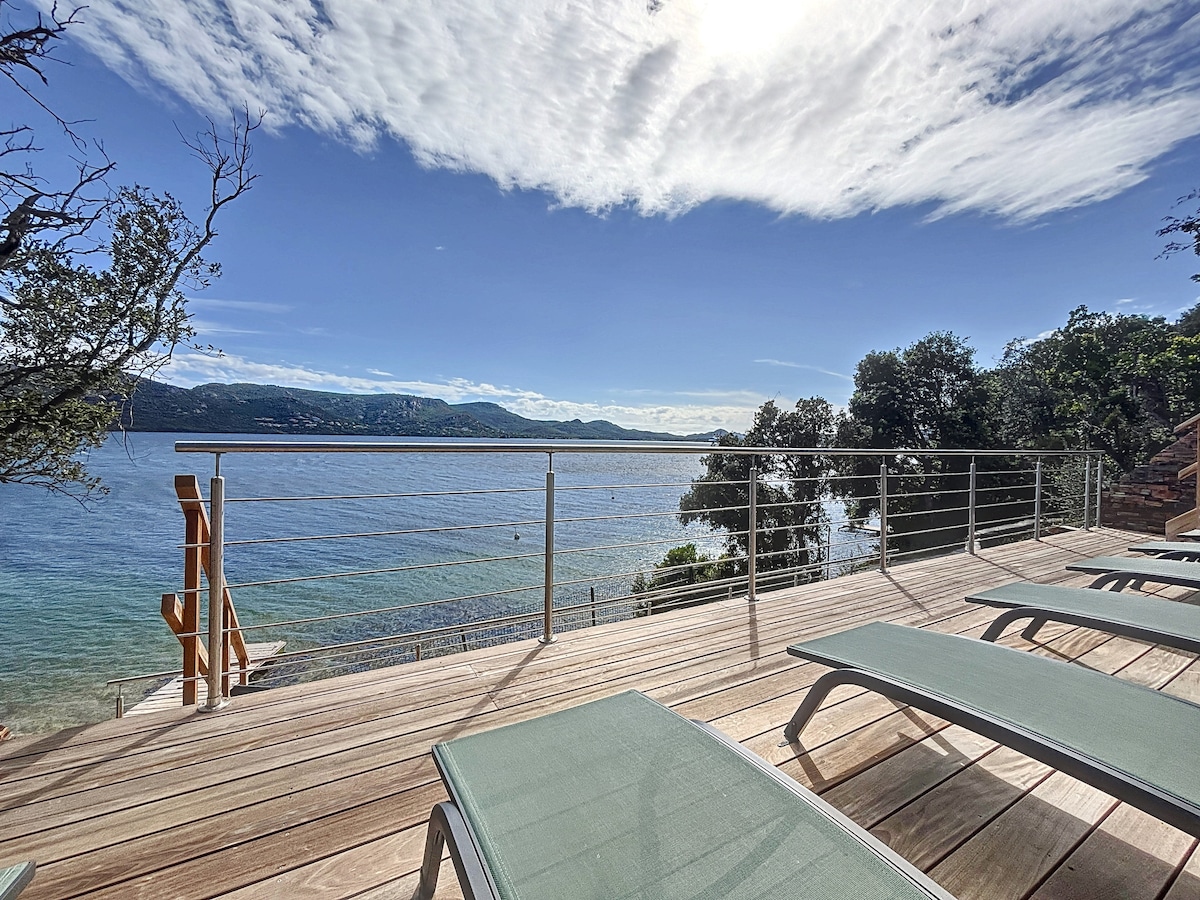 A deck overlooking the serene ocean features several lounge chairs placed for relaxation. The calm water reflects the sky, with gentle waves lapping at the shore, while the distant hills provide a scenic backdrop under a partly cloudy sky.