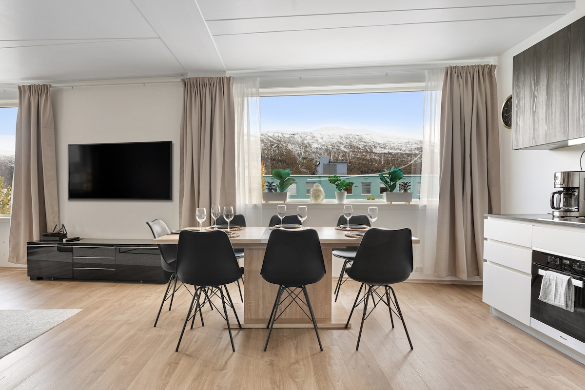 A modern living space is featured, showcasing a dining table surrounded by six black chairs. A large window offers a view of mountains, with green plants adorning the windowsill. A flat-screen TV is mounted on the wall, and kitchen appliances are neatly arranged.