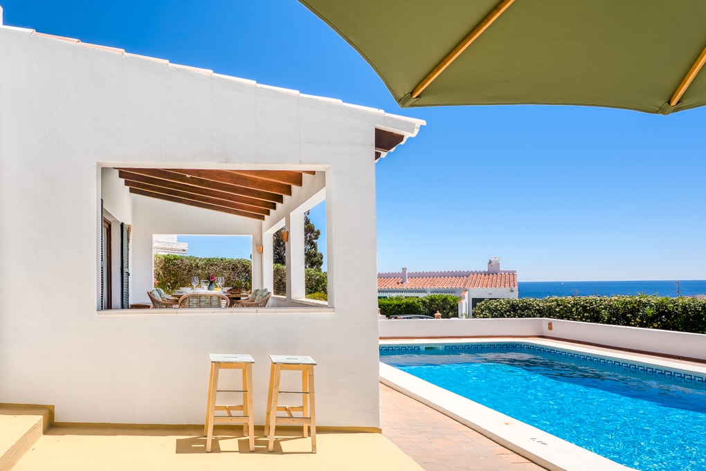An outdoor view showcases a private pool flanked by hedges, with bright blue water reflecting the clear sky. In the background, a spacious terrace is visible through an open window, inviting natural light into the space. Two stools are positioned beside the pool.