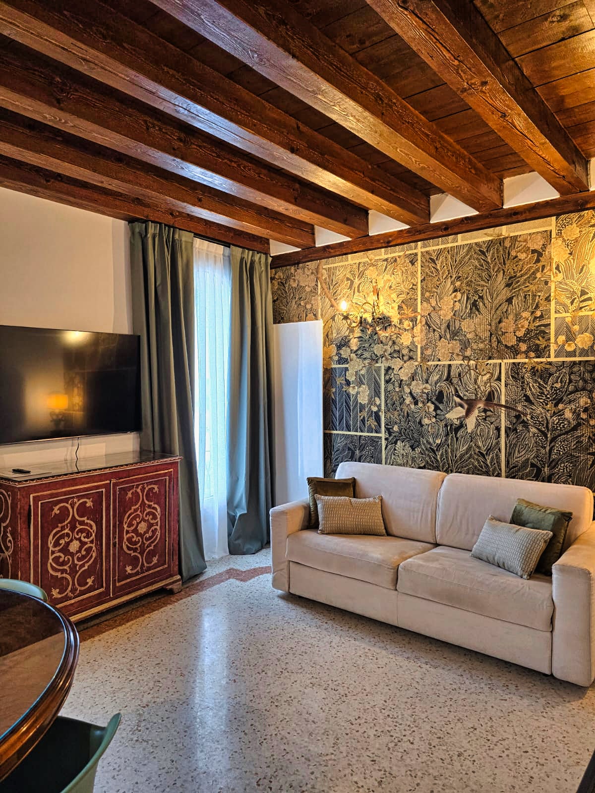 The living area features exposed wooden beams and artistic wall coverings that evoke nature. A comfortable sofa in light tones is positioned near a flat-screen TV, with accent pillows enhancing the space. Natural light filters through the curtain-draped window, creating a welcoming environment.