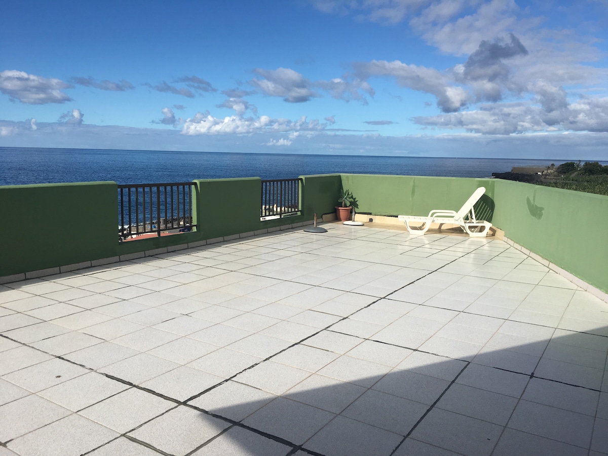 A spacious terrace features panoramic ocean views, with a sectional railing for safety. Two lounge chairs are positioned on the tiled surface, inviting relaxation. The sky is partly cloudy, creating a serene environment that enhances the overall tranquility of the area.