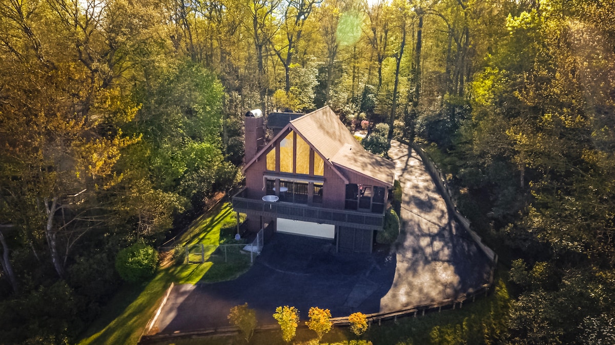 An aerial view of a spacious mountain-top vacation house is displayed, set against a backdrop of vibrant autumn foliage. The house features a distinct tan roof and large windows, with a paved driveway leading to the entrance, surrounded by greenery.