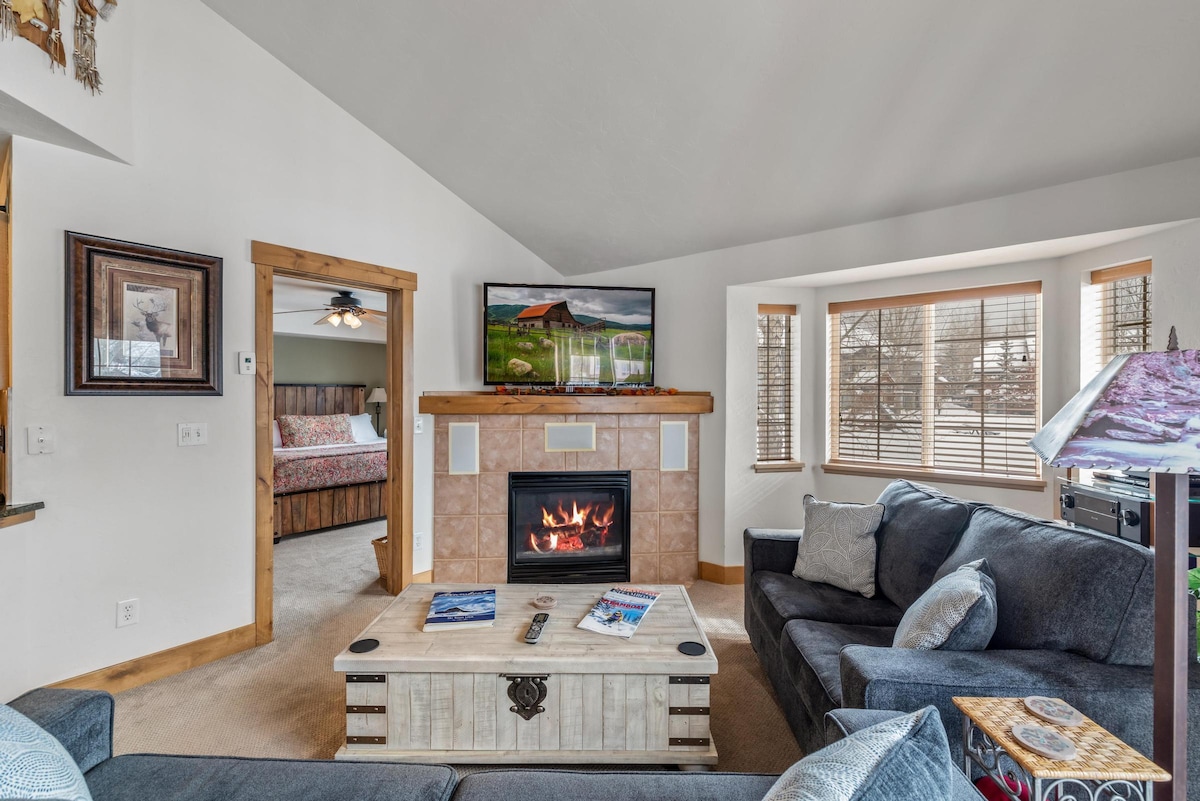 A spacious living area features a comfortable dark sofa, arranged around a coffee table with various magazines. A gas log fireplace is positioned beneath a mounted television. Large windows provide natural light, with views of the surrounding scenery, enhancing the cozy ambiance.