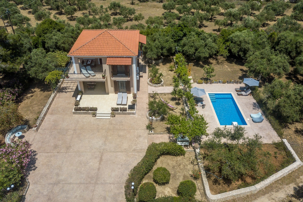 Aerial view of a two-storey house surrounded by greenery. The property features a swimming pool with sun loungers, a spacious patio with outdoor furniture, and well-maintained gardens, all set in a tranquil area. A pathway leads to the house from the pool area.