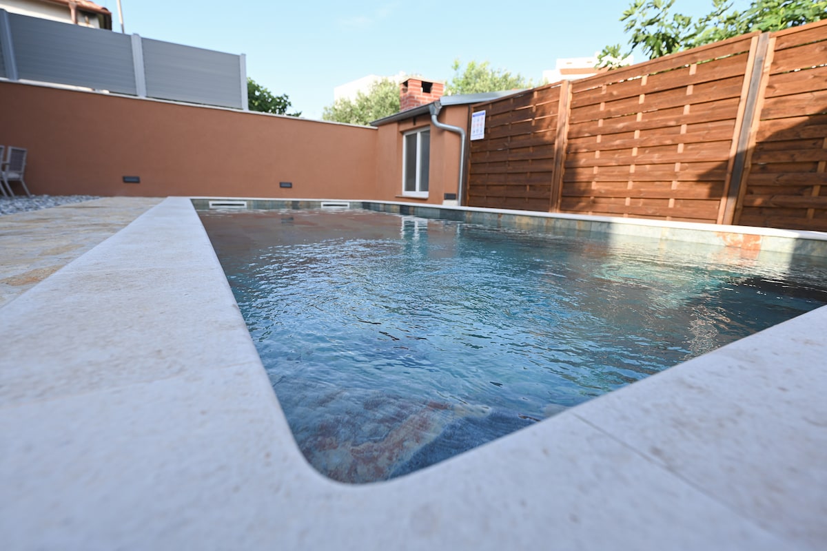 A heated swimming pool is framed by a smooth, light-colored stone edge. The clear water reflects sunlight, surrounded by an enclosed area featuring wooden fencing and greenery. A few outdoor chairs are positioned nearby, offering a relaxing setting.