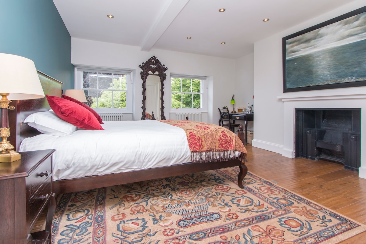 A private room features a king-size bed with crisp white linens and a decorative throw, positioned against a green accent wall. Natural light fills the space through large windows, and a vintage-style mirror adds character. A small workspace and fireplace are also visible.