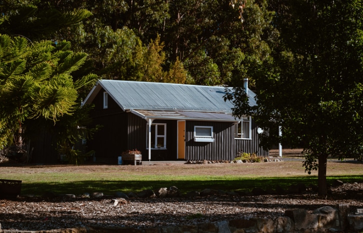 A charming waterfront cottage is set against a backdrop of lush greenery. Its dark exterior contrasts with the light-colored windows and door, while gravel pathways lead to the entrance. A tranquil outdoor area features a mix of trees and well-maintained grounds.
