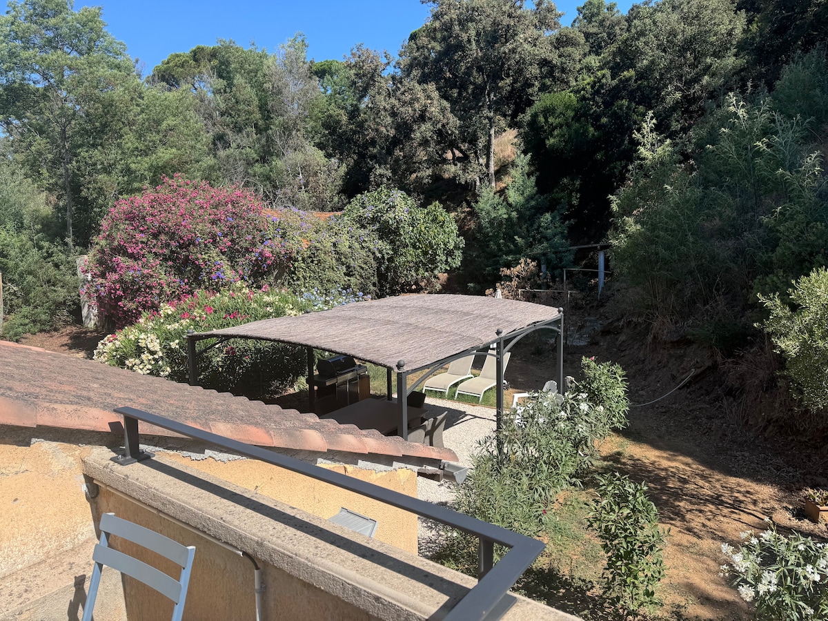 A view from above reveals a shaded outdoor area with a canopy, surrounded by lush greenery and vibrant flowers. The natural landscaping creates a serene environment, emphasizing the connection between the space and nature.