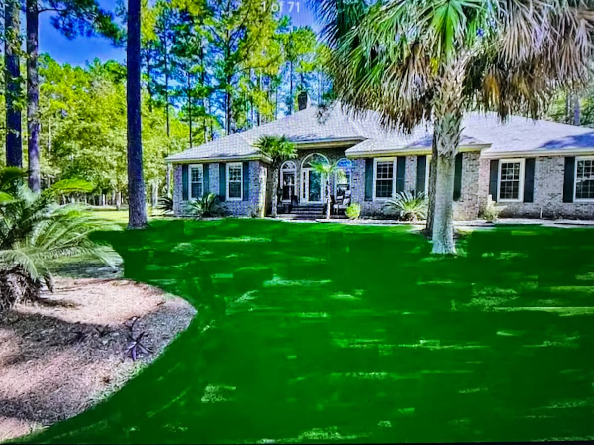The exterior of a spacious home is presented, surrounded by lush greenery and tall trees. The welcoming entrance features architectural details with large windows, allowing natural light throughout the interior. The well-maintained yard enhances the serene atmosphere.