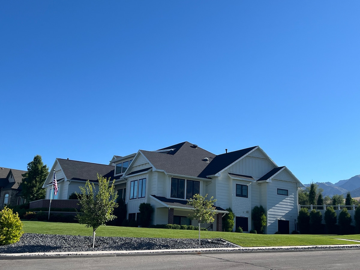 The exterior of a modern two-story home features a light-colored façade with dark roofing. Lush greenery surrounds the property, including several trees and well-manicured shrubs. A clear blue sky provides a bright backdrop to this spacious residence.