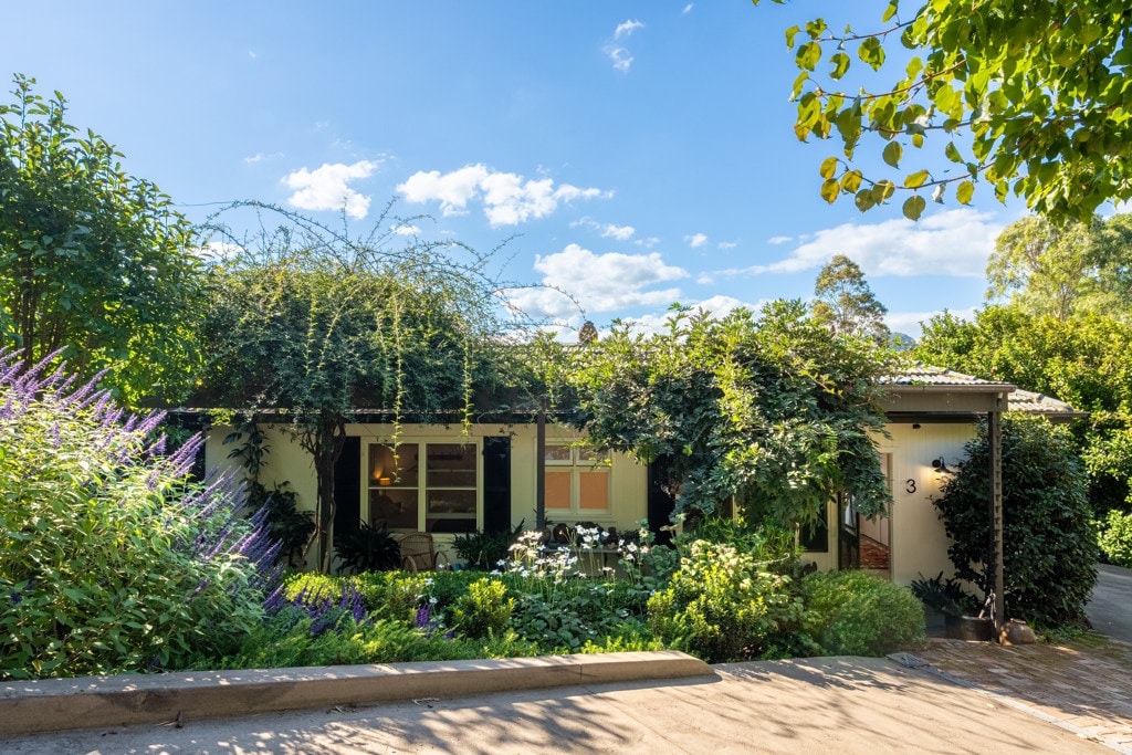 The entry of a charming timber cottage is surrounded by lush greenery and colorful flowering plants. A stone pathway leads to the front door, complemented by a vibrant blue sky and scattered clouds overhead.