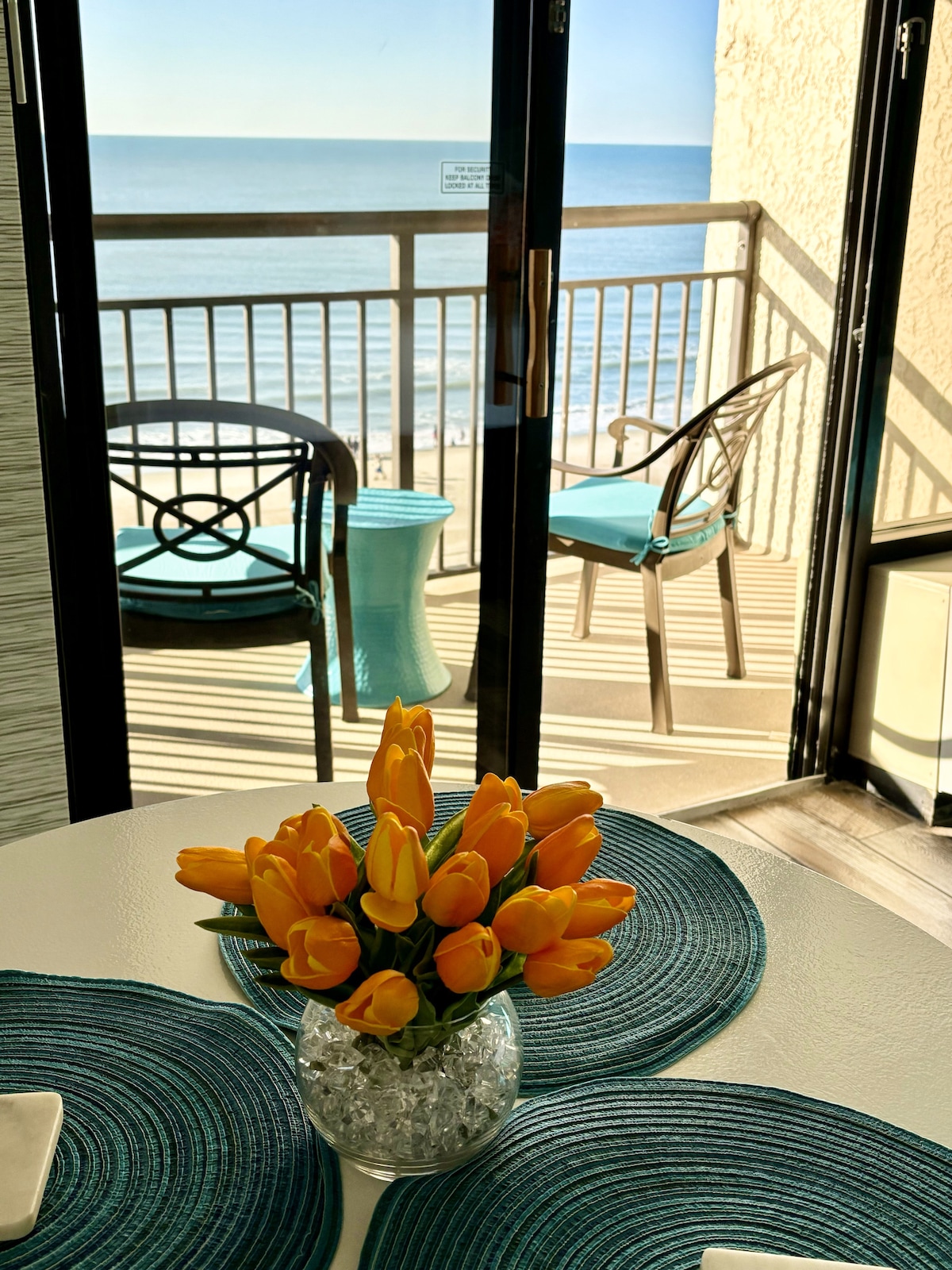A bright dining area features a white table adorned with a vase of orange tulips. Floor-to-ceiling glass doors open to the private balcony, revealing ocean views. Two outdoor chairs and a side table are seen, complementing the coastal setting.