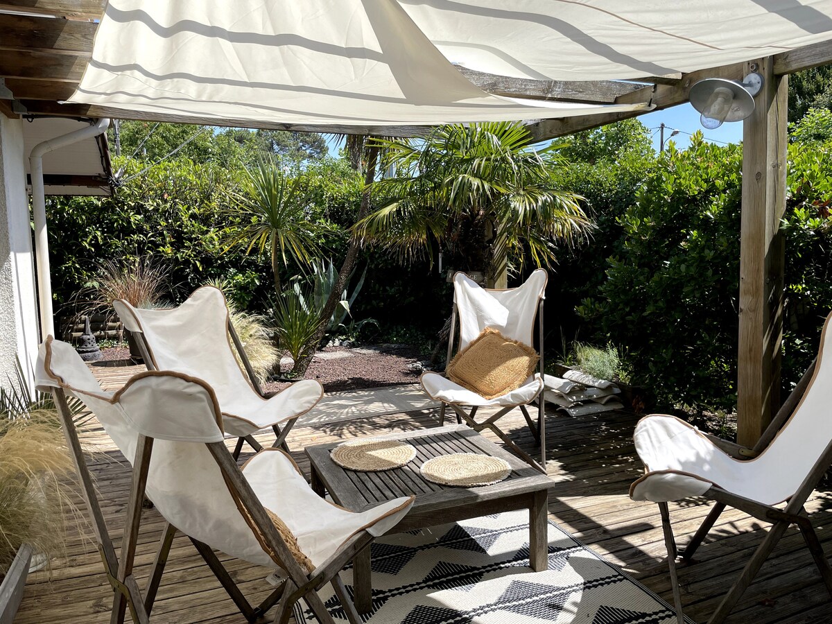 A spacious outdoor terrace features several light-colored chairs arranged around a wooden table. A canopy offers shade, while greenery forms a lush backdrop, adding to the tranquil setting. Natural textures are present through wooden surfaces and woven table placemats.