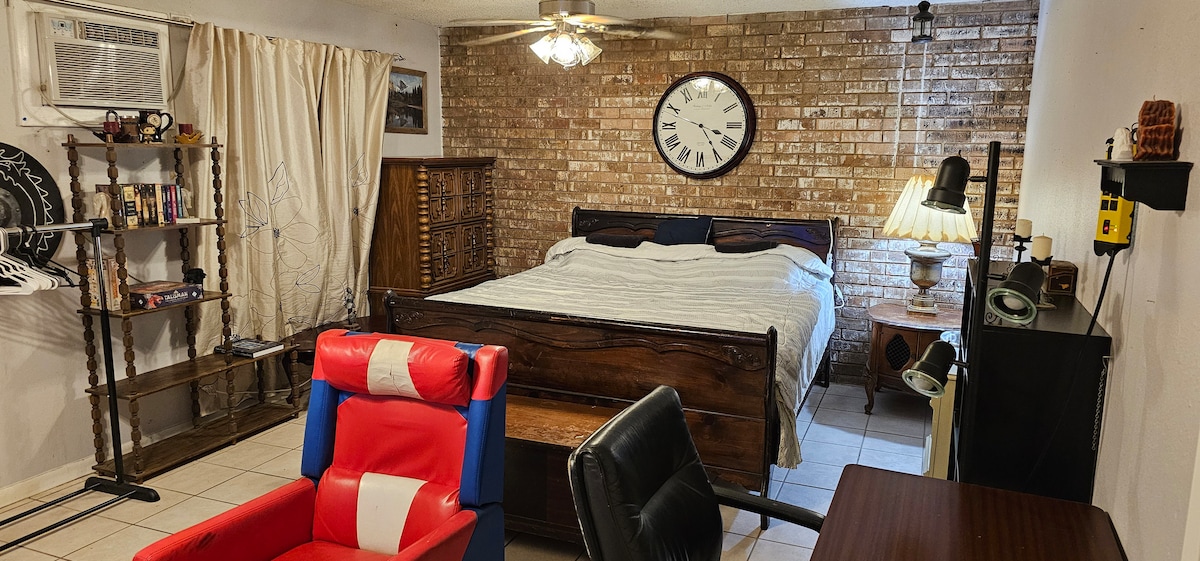 The spacious room features a king size bed positioned against a brick wall, complemented by a large wall clock. A comfortable chair and desk are visible, alongside a lamp and shelving unit holding decorative items. Natural light filters through the curtain-covered window.