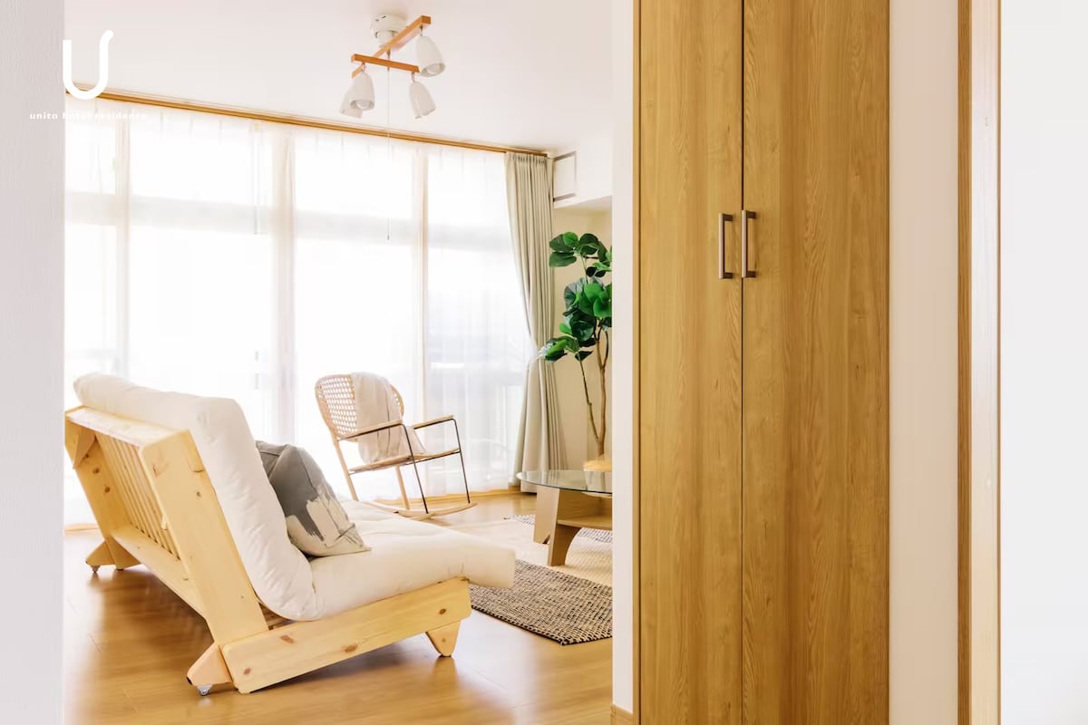 A bright living area is seen, featuring large windows that allow natural light to fill the space. A wooden folding lounge chair and a rattan chair provide seating options. A neutral-colored area rug lays on the floor, contributing to a relaxed atmosphere.