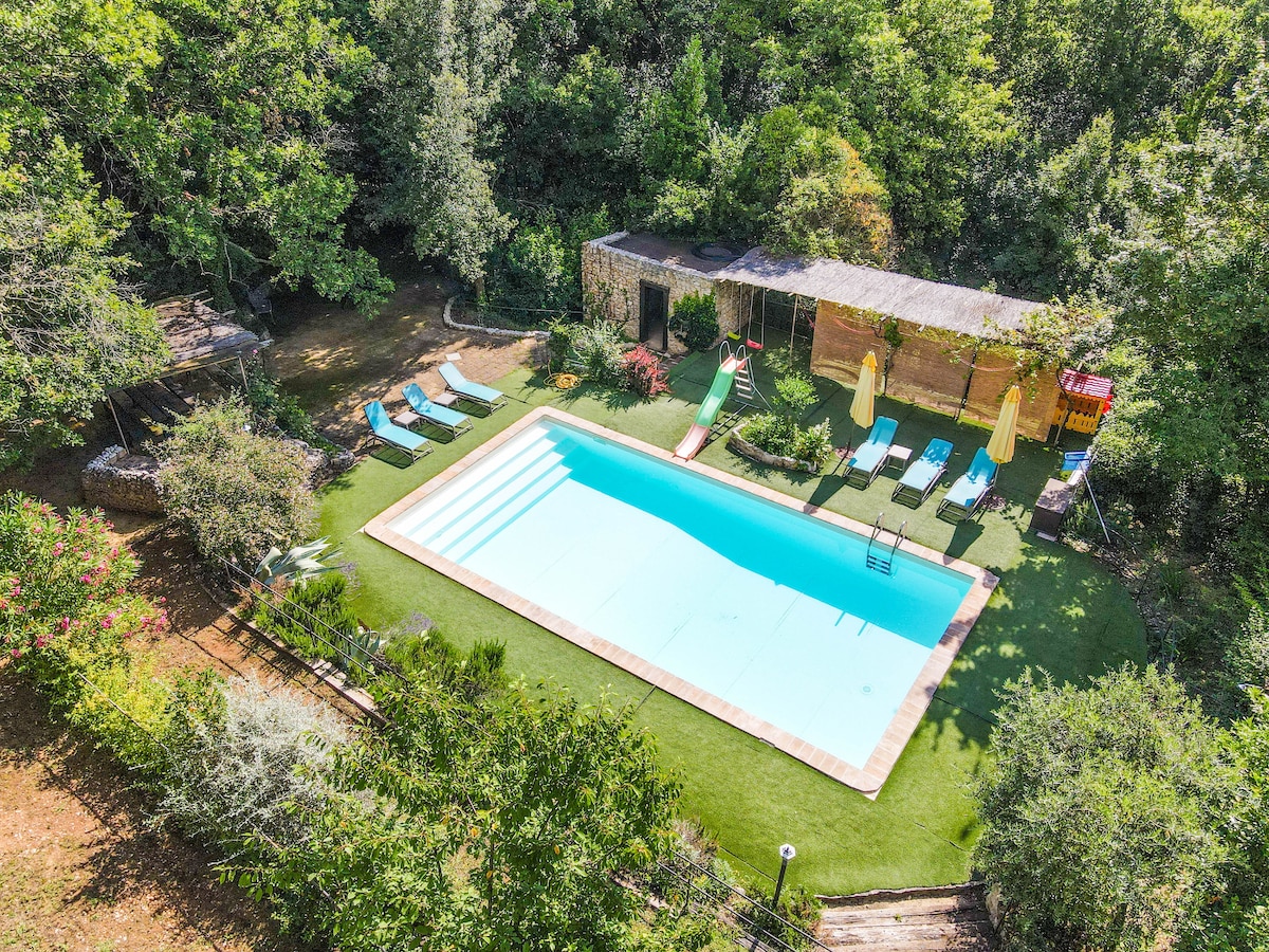 An aerial view of the private garden features a spacious pool surrounded by sun loungers and umbrellas. The pool is neatly bordered by grass, with a wooden structure nearby that includes changing facilities. Lush greenery encircles the area, offering a serene outdoor setting.