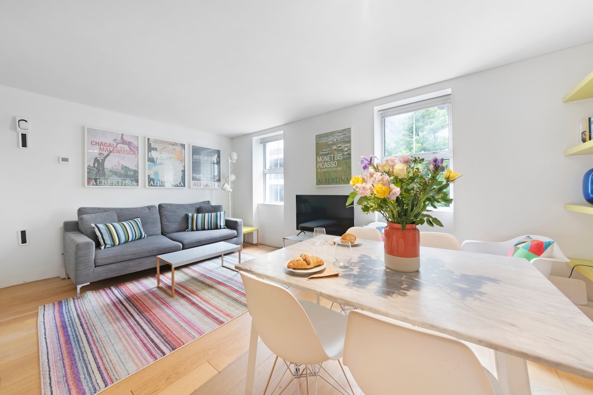 A modern living space features a grey sofa with colorful cushions, accompanied by a marble dining table adorned with a vase of fresh flowers. Natural light floods in through two windows, illuminating a multi-colored rug that adds warmth to the room, while a TV is positioned nearby.