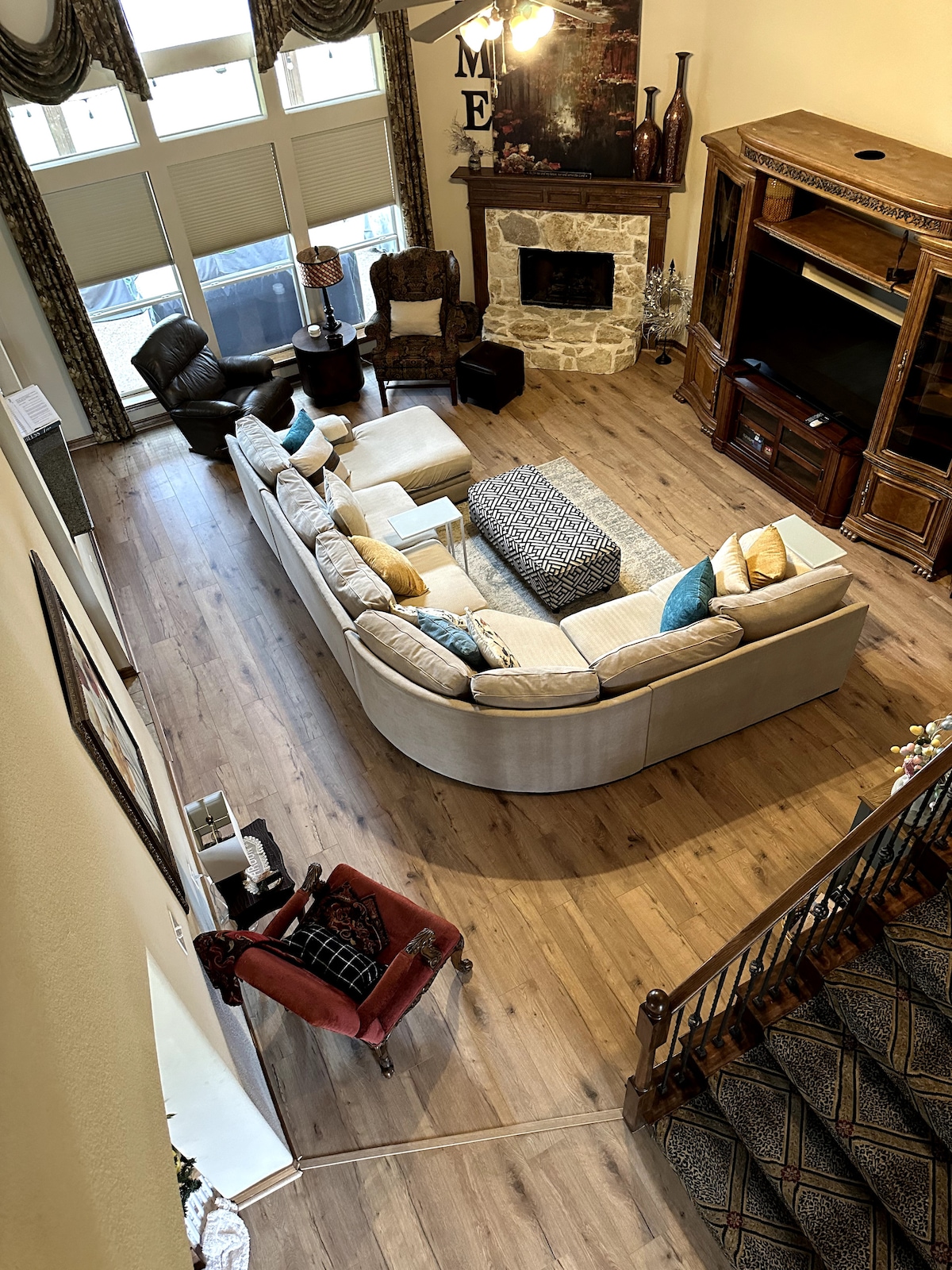 An open family room is viewed from above, displaying a spacious sectional sofa arranged around a coffee table. Natural light enters through large windows, highlighting the stone fireplace and wooden entertainment unit. A cozy armchair and a decorative vase add character to the space.