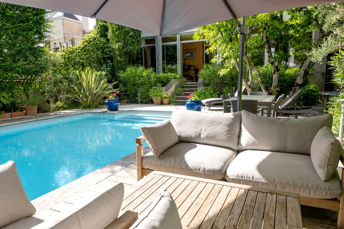 An outdoor seating area features soft grey cushions on a wooden sofa, overlooking a private pool surrounded by lush greenery. Potted plants and blue ceramic accents enhance the tranquil ambiance of the garden space, inviting relaxation by the water.