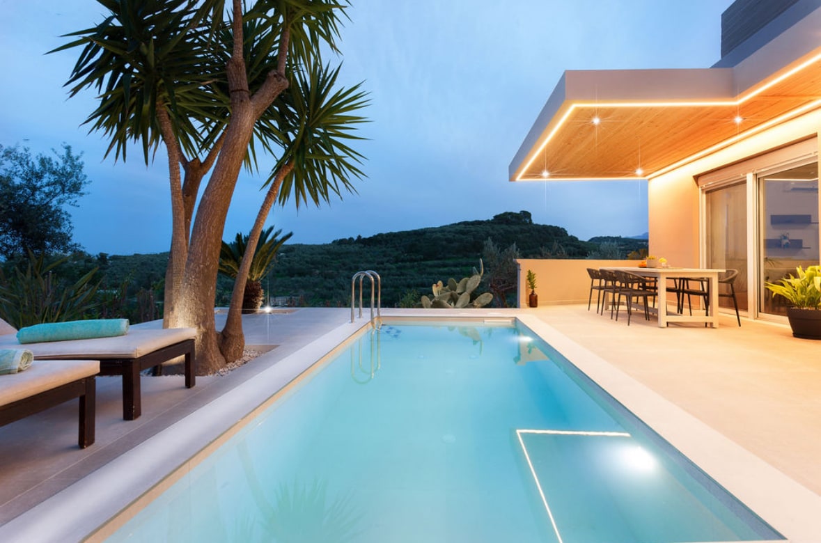 An outdoor terrace features a heated swimming pool surrounded by lounge chairs and a dining area. Palm trees and landscaped greenery provide a natural backdrop, while subtle lighting enhances the evening ambiance. Views of the rolling hills can be seen in the distance.