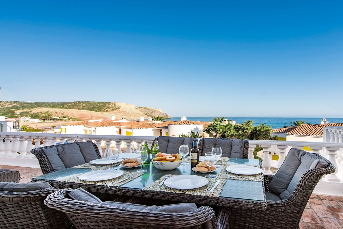 An outdoor dining area features a large woven table surrounded by cushioned chairs. A table setting includes plates, glasses, and fresh fruit, with the ocean and hills visible in the background under a clear blue sky.