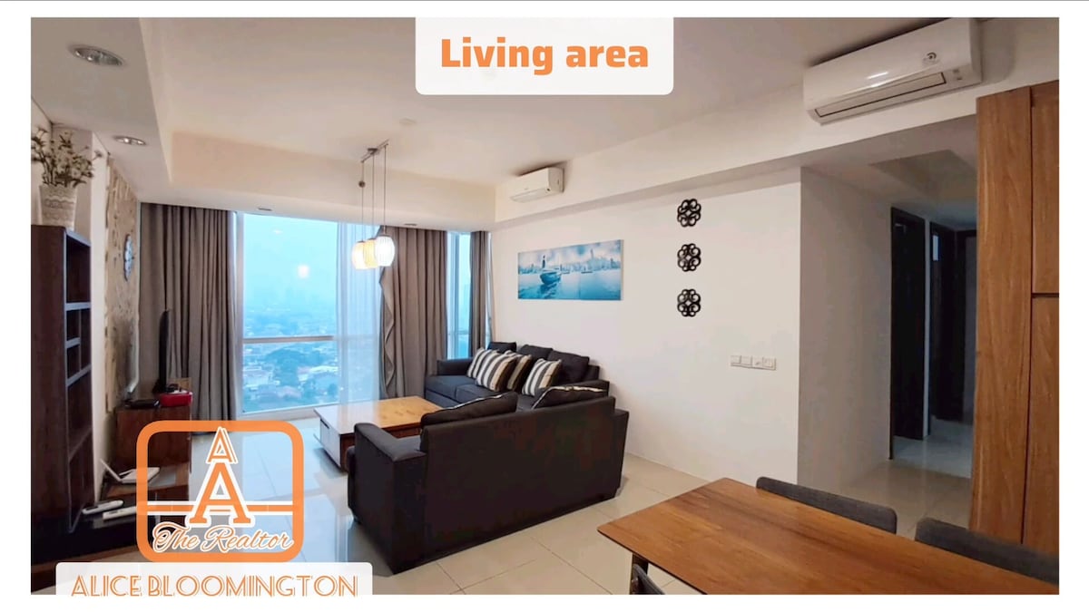 The living area features a spacious layout with a comfortable seating arrangement including a dark sofa and a coffee table. Floor-to-ceiling windows allow natural light and offer views of the surrounding area. A dining table is positioned nearby, creating a cohesive living space.