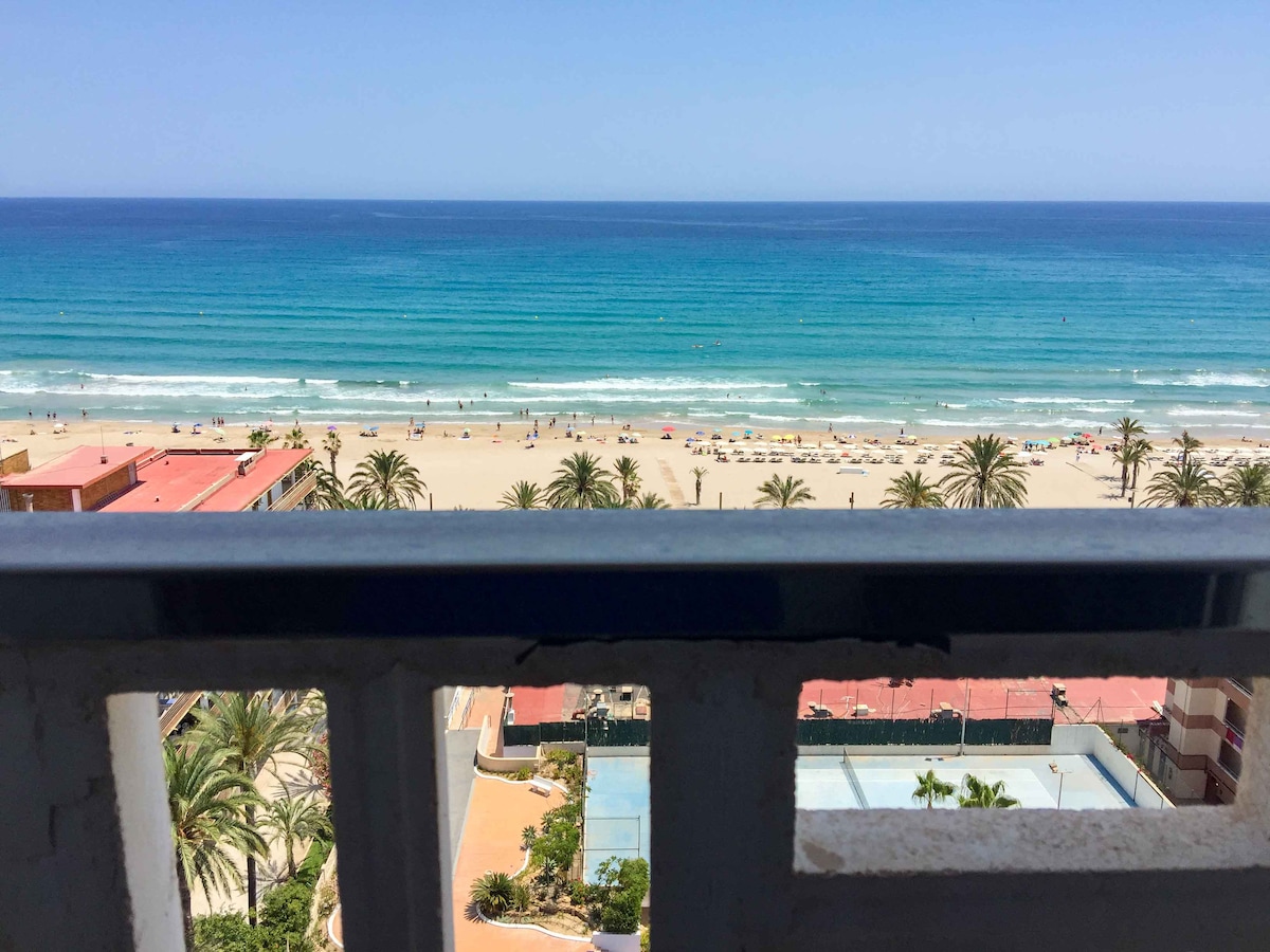 A panoramic view of the beach is displayed from a balcony railing, showcasing clear blue waters and gentle waves. Sun loungers and umbrellas are arranged along the sandy shore, while palm trees line the beach area, contributing to a vibrant coastal setting.