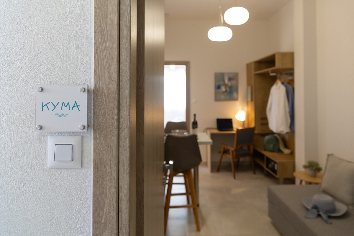 An inviting entrance is highlighted by a sign reading 'KYMMA'. A bright, well-furnished living area is visible beyond, featuring a dining table and chairs, along with a small workspace. Light fixtures suspended from the ceiling create a warm ambiance.