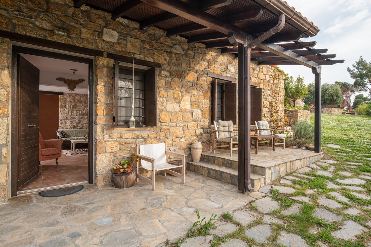 A stone cottage's front porch features wooden beams and natural stone flooring. Comfortable outdoor seating is arranged under the cover, with planters adding greenery. The garden space beyond is visible, showcasing a well-maintained lawn leading to trees.