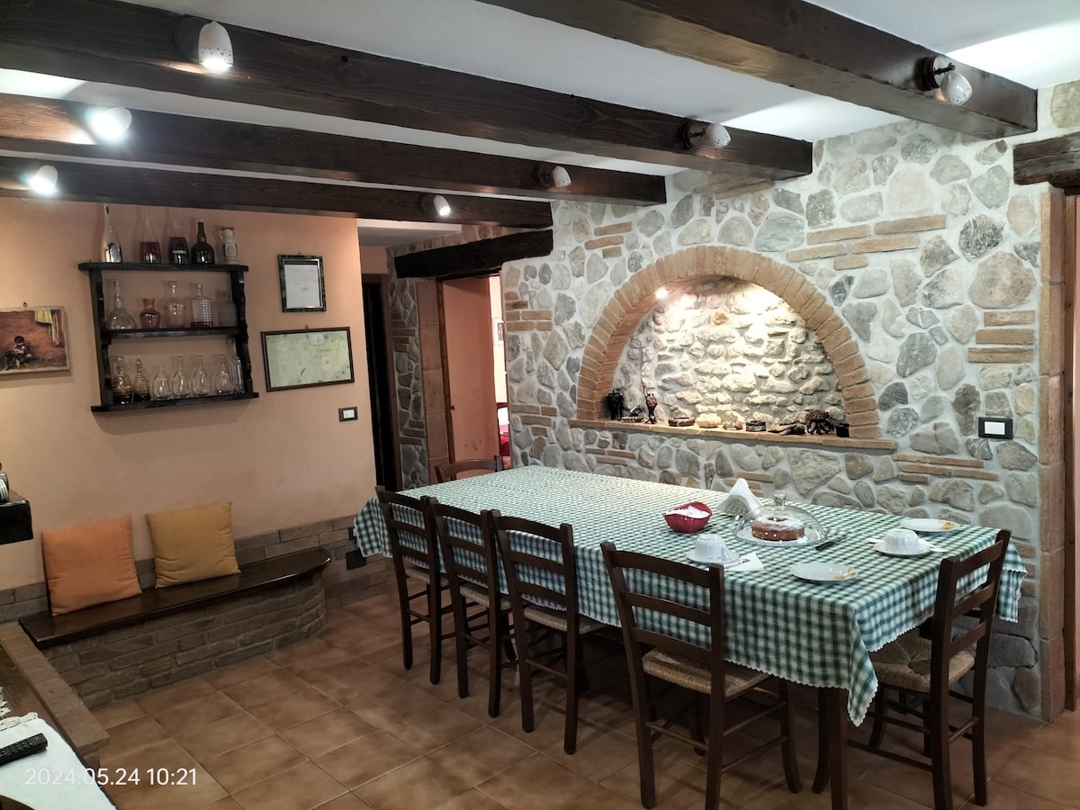 A rustic dining area features a large wooden table surrounded by chairs, set with plates and cutlery. A brick archway reveals a stone wall and a decorative fireplace. Warm lighting highlights the space, creating a welcoming environment for gatherings.