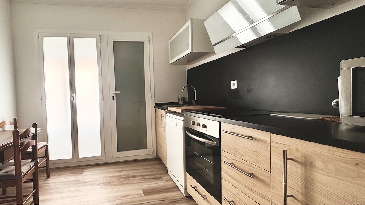 A modern kitchen is equipped with wooden cabinetry and sleek black countertops. Large windows allow natural light to fill the space. Essential appliances, including an oven, microwave, and sink, are integrated into the design. A small dining area with wooden chairs is visible.