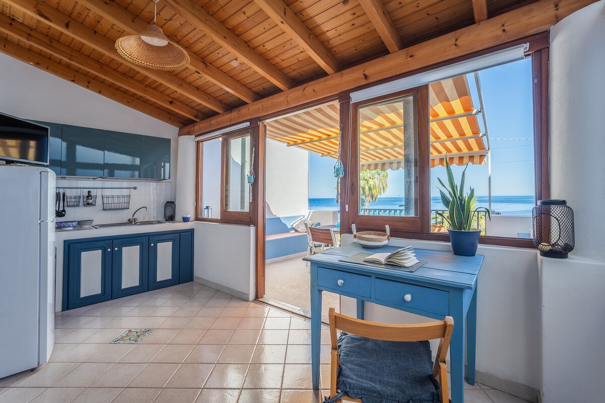 An open kitchen area features blue cabinetry and a white refrigerator. Large windows offer views of the sea, while light filters through striped awnings onto a cozy patio space. A simple wooden table and chair create a welcoming study area beside the kitchen.
