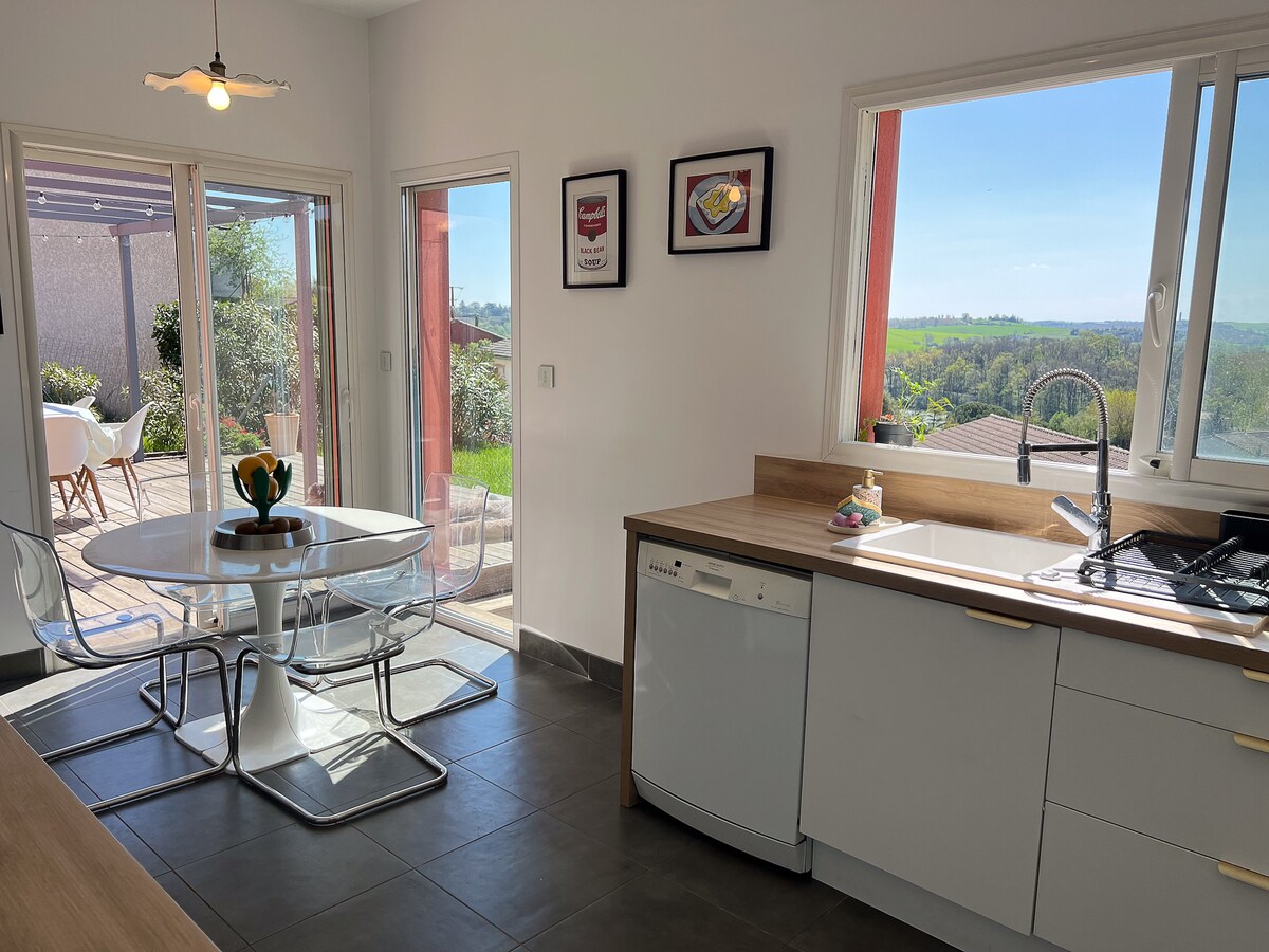 A modern kitchen is depicted with natural light streaming through large glass doors that open to an outdoor terrace. A round table with transparent chairs is positioned next to the window, while a sleek countertop features a dishwasher and a sink overlooking the serene landscape.