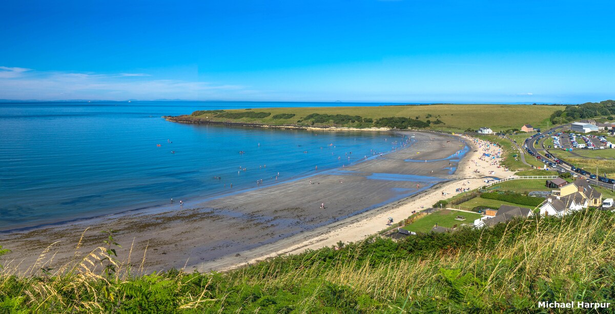 A panoramic view of a serene beach, bordered by gentle hills and lush greenery. Beachgoers can be seen enjoying the calm waters, while sandy areas are visible along the shoreline. Nearby, a cluster of colorful beach huts and parked cars is observed.