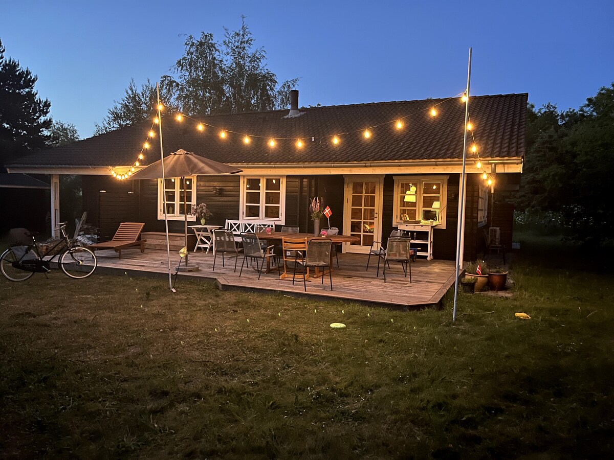 The exterior of the summer house is shown at dusk, enhanced by warm string lights that illuminate the patio area. Various seating options, including wooden loungers and dining tables, are arranged on a wooden deck. Lush greenery surrounds the house.