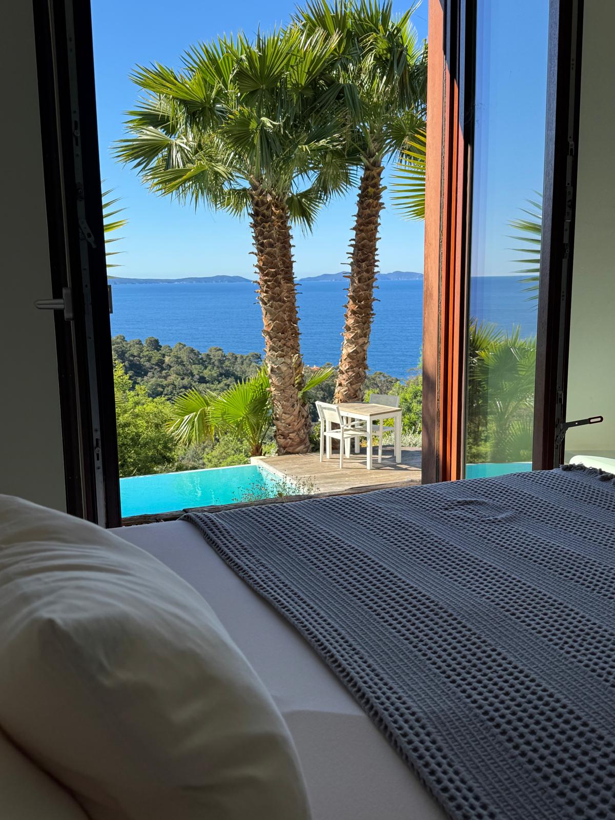 A serene view is offered from the bedroom, featuring palm trees framing the landscape. The azure sea can be seen in the distance, with a glimpse of a designated seating area next to a refreshing infinity pool.