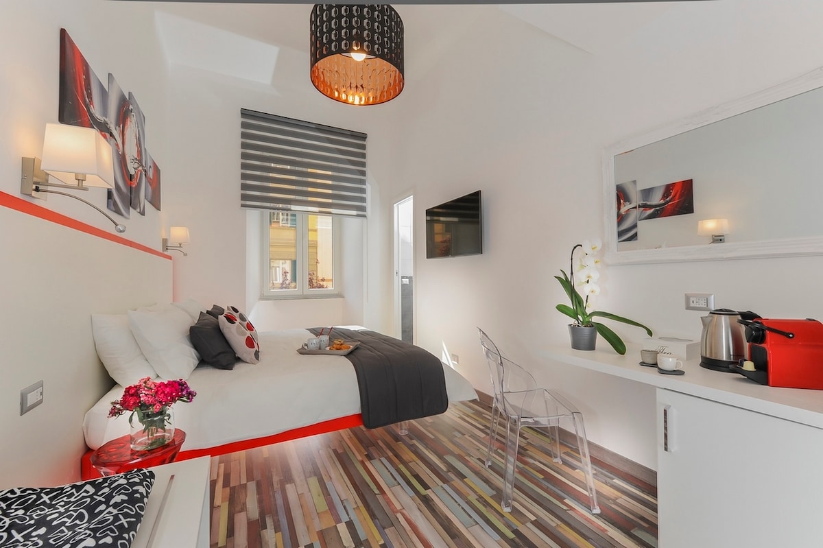 A well-lit guest room features a spacious bed with a gray throw and decorative pillows. A flat-screen TV is mounted on the wall. A small table holds a coffee machine and kettle, while a potted plant adds a natural touch. Decorative art pieces complement the modern decor.