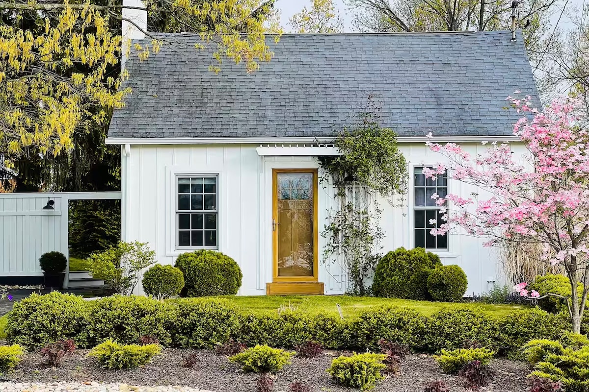 A charming cottage exterior is showcased, featuring a light-colored facade complemented by well-manicured shrubs and flowers. A wooden front door invites entry, while pink blossoms on a nearby tree add a touch of color to the serene landscape.