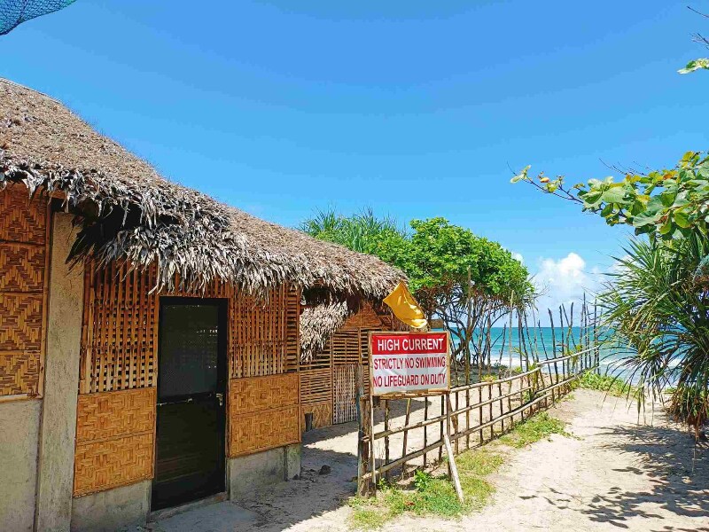 A charming native wooden house with a thatched roof stands beside the beach. A bright warning sign about high currents is prominently displayed, while lush greenery and the ocean can be seen in the background under a clear blue sky.